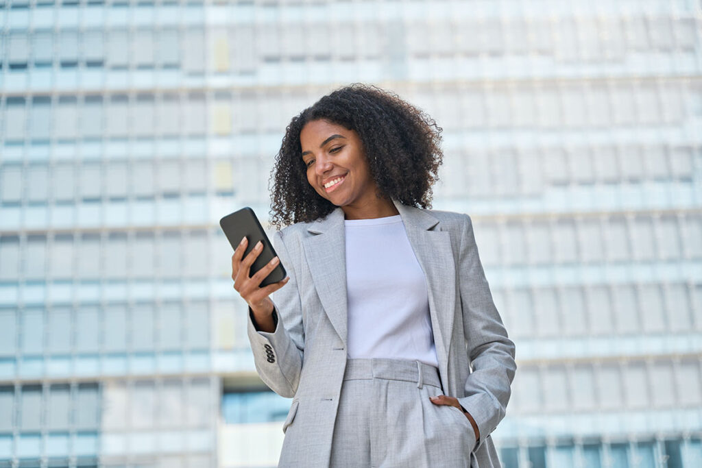 Smiling lady professional holding smartphone standing at city street building looking at mobile working outside office.