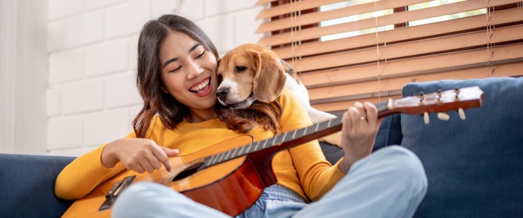 Young female owner feeling happy and relax, enjoy play guitar and spend leisure time with her lovely pet puppy together in house